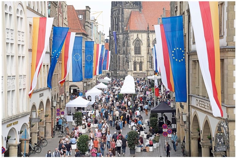 Comeback für Münsters Hansemahl auf dem Prinzipalmarkt: Die Giebelhäuser sind geschmückt mit gelb-rot-weißen Münsterfahnen und blauen Europabannern, eine lange Tafel und ein Loungebereich luden zum Platznehmen ein, und die Kaufleute bedienen ihre Gäste.
