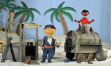 Die Marionetten Scheinriese Tutur, Lukas und Jim Knopf stehen im Puppenkistenmuseum mit der zerstörten Lokomotive Emma in einem Wüstenszenario.
